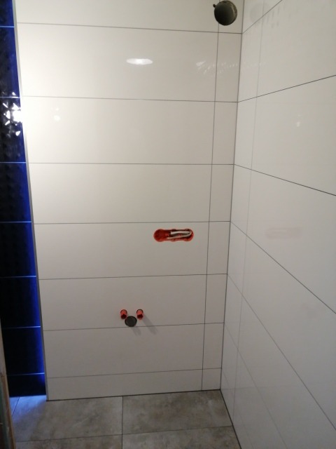 Ściana łazienki wyłożona białymi, podłużnymi płytkami z widocznymi otworami na armaturę i rurami instalacyjnymi w kolorze czerwonym, z fragmentem dekoracyjnej czarnej ściany po lewej stronie.