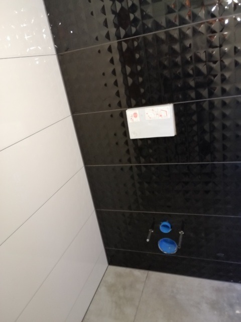 Fragment ściany łazienki z czarno-białymi płytkami, widoczne przyłącza hydrauliczne i przycisk spłuczki w zabudowie podtynkowej.