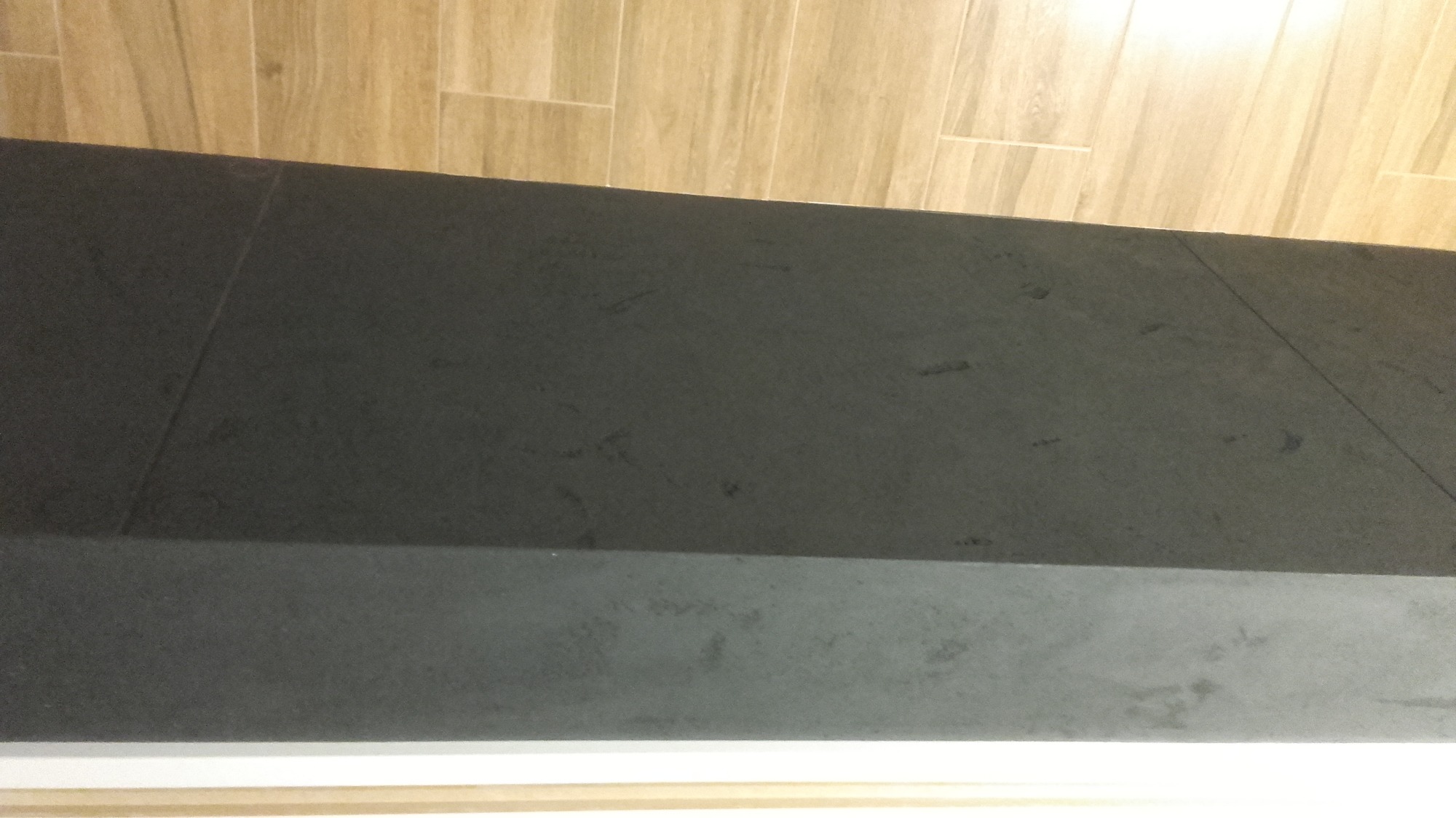 Fragment ściany wykończonej ciemnoszarą płytą z delikatną fakturą, widoczne połączenia płyt, górą jasne płytki imitujące drewno.