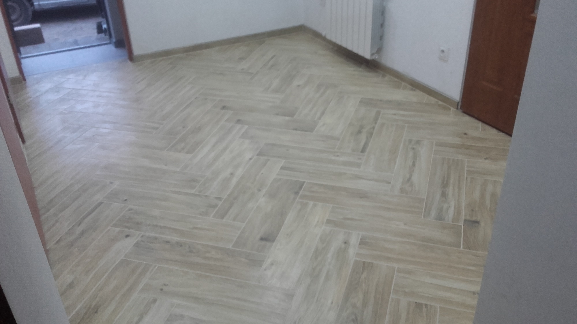 Ułożona w jodełkę podłoga z płytek imitujących drewno w jasnym odcieniu, widoczne fragmenty ścian, drzwi i grzejnika w odnowionym pomieszczeniu.