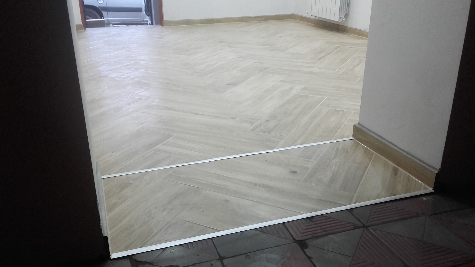 Jasne, drewnopodobne płytki ułożone w jodełkę na podłodze, z metalową listwą progową oddzielającą je od ciemniejszych płytek w innym pomieszczeniu, widok z perspektywy przejścia.