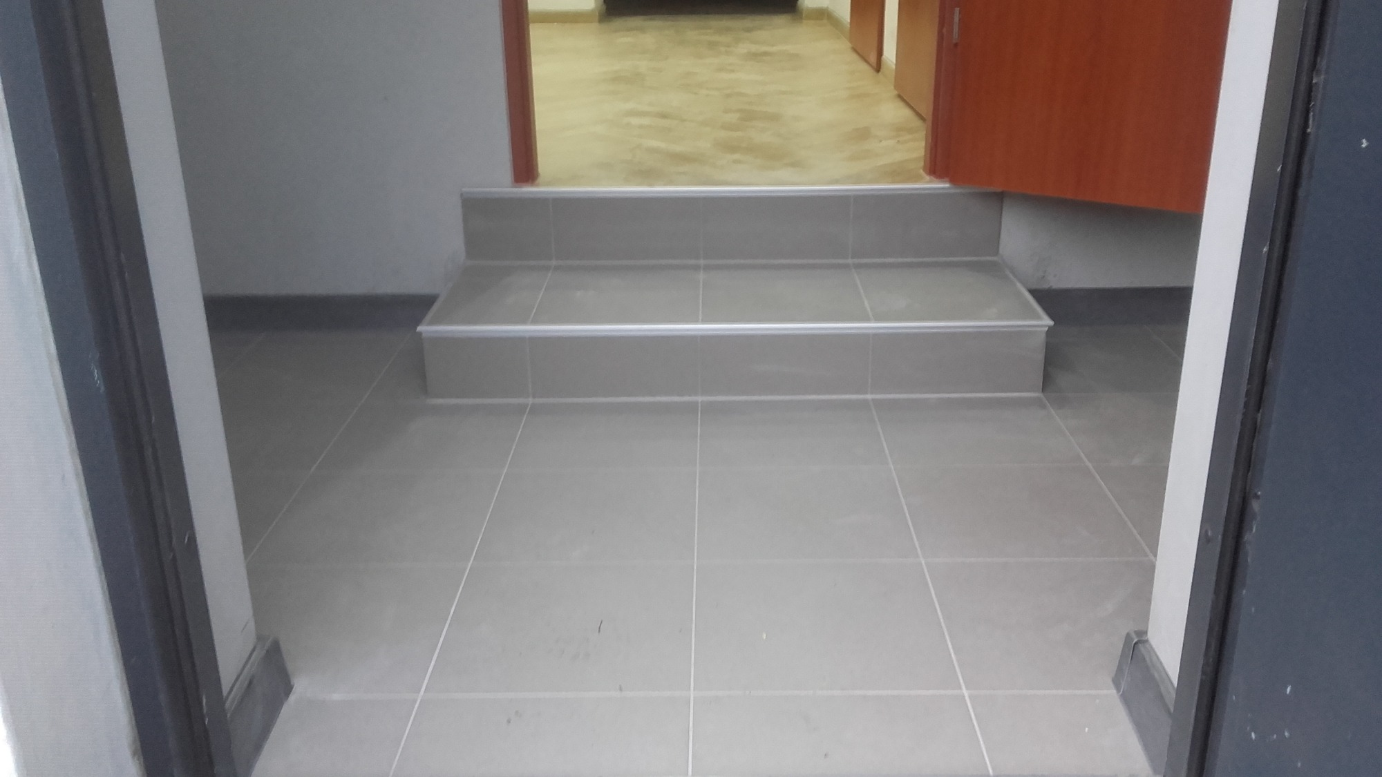 Wykończone schody wejściowe do pomieszczenia, wyłożone szarymi płytkami ceramicznymi z aluminiowymi listwami krawędziowymi, widoczne z poziomu podłogi.