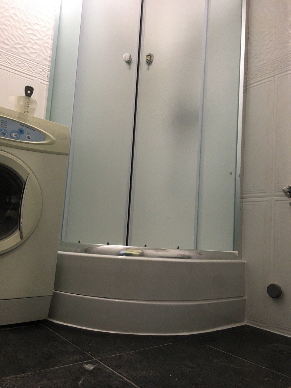 Narożna kabina prysznicowa z matowego szkła w łazience z ciemną podłogą i białą pralką obok.