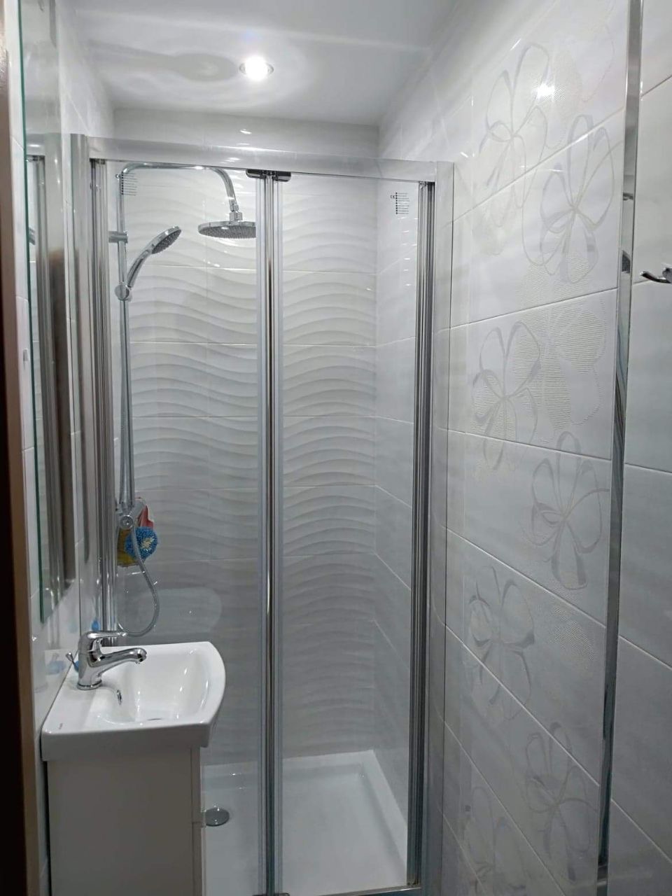 Nowoczesna, mała łazienka z kabiną prysznicową ze srebrnymi ramami, białą umywalką z szafką i płytkami z delikatnym wzorem kwiatowym na ścianach.