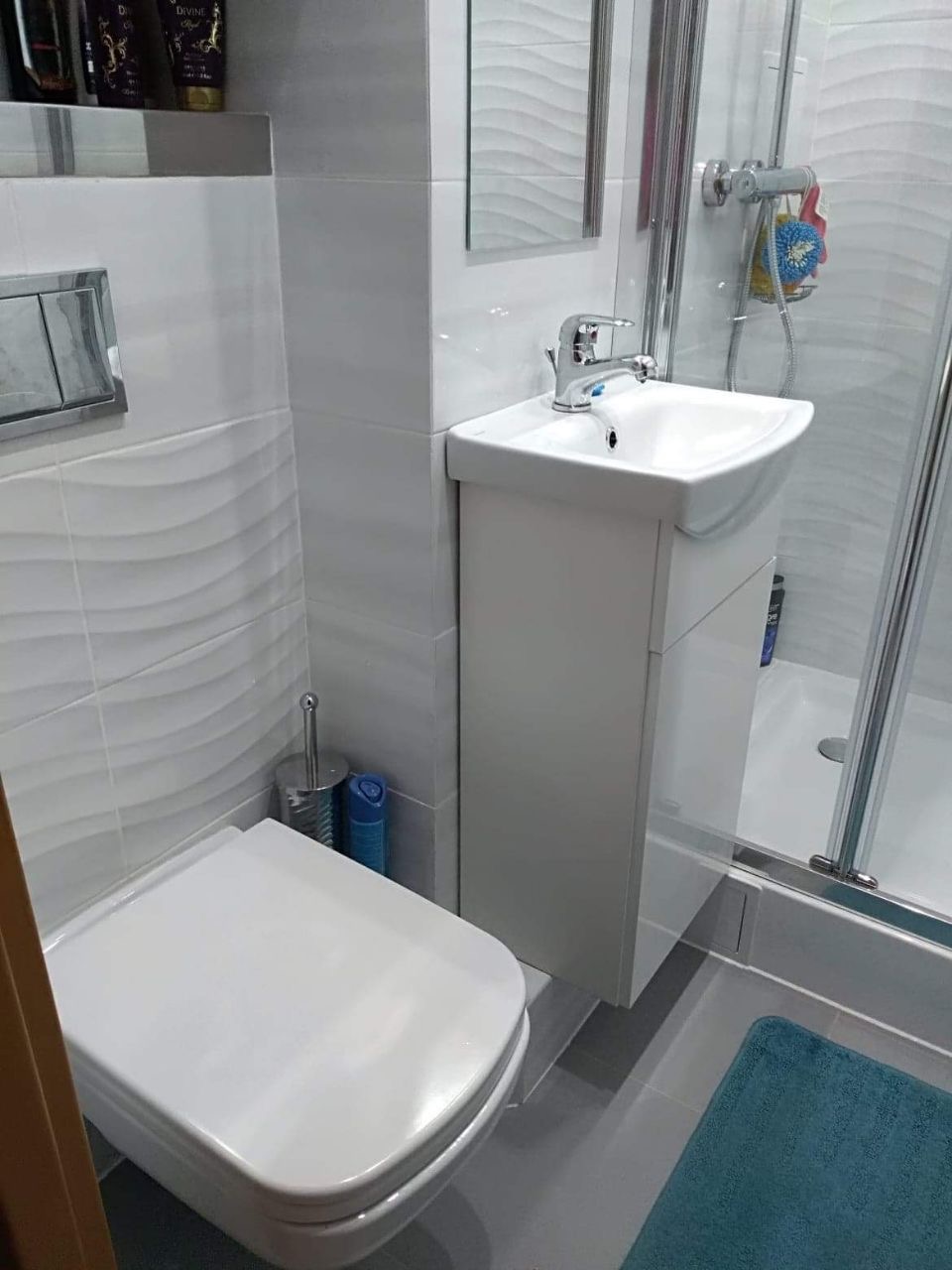 Nowoczesna, mała łazienka z białą armaturą i płytkami o falistej strukturze, kabina prysznicowa ze srebrnymi profilami, niebieski dywanik podłogowy.