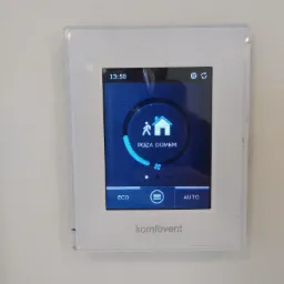 Biały panel sterowania rekuperacją firmy Komfovent zamontowany na ścianie, wyświetlający tryb 'Poza domem' na niebieskim ekranie.