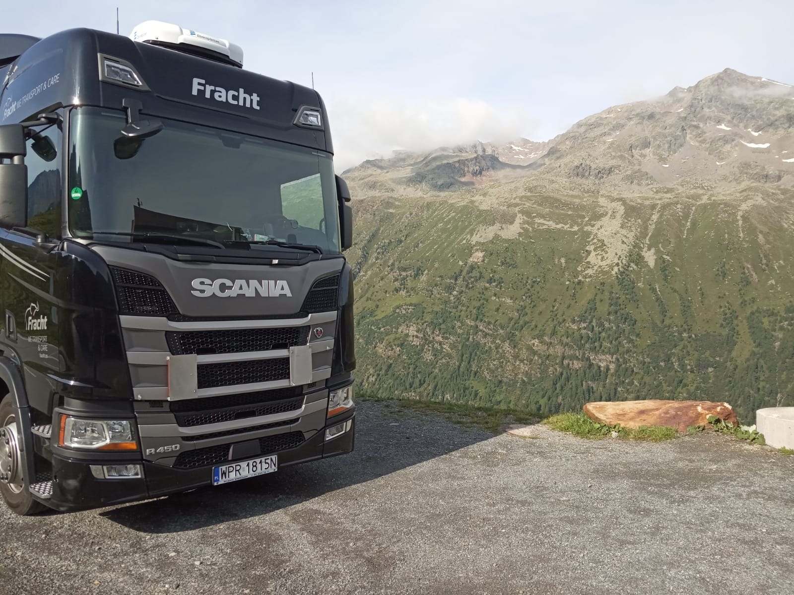 Czarna ciężarówka Scania firmy Fracht zaparkowana na żwirowym parkingu z widokiem na górzysty krajobraz w tle.