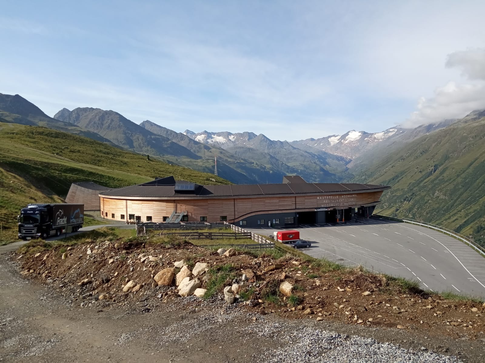 Czarna ciężarówka z naczepą firmy Fracht na tle górskiej scenerii z budynkiem Mautstelle - Hoengurgl, Restaurant z. Team, Motorcycle Museum.