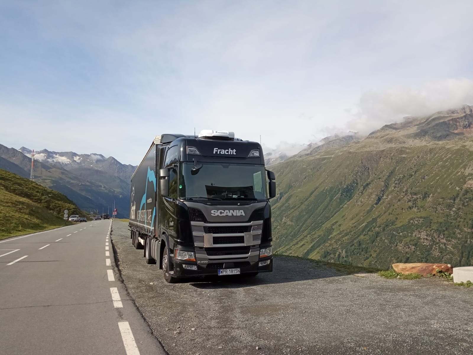 Czarna ciężarówka Scania Fracht z naczepą na tle górskiego krajobrazu z widoczną drogą i innymi pojazdami w oddali.