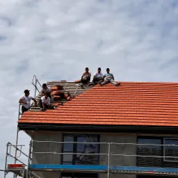 Grupa dekarzy układa czerwoną dachówkę na nowym dachu domu w trakcie budowy, widoczne rusztowania i zabezpieczone okna.