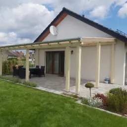 Drewniana pergola z płaskim dachem nad tarasem z kostki brukowej przy domu jednorodzinnym z ciemną dachówką, widok na ogród z zadbanym trawnikiem i rabatami kwiatowymi.