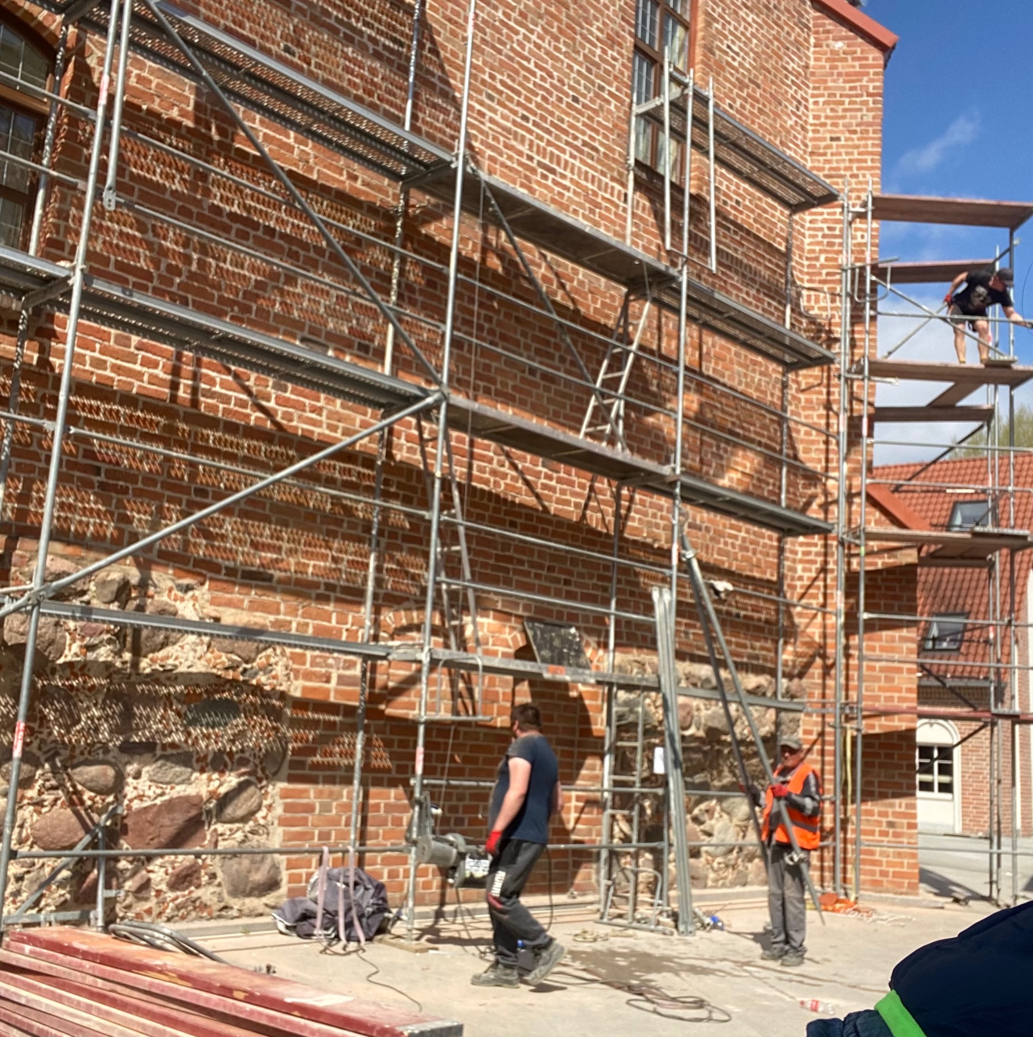 Pracownicy na rusztowaniu przy ceglanym budynku z kamienną podstawą, jeden z nich pracuje na wysokości, drugi w kamizelce odblaskowej stoi na ziemi, w tle czerwony dach innego budynku.