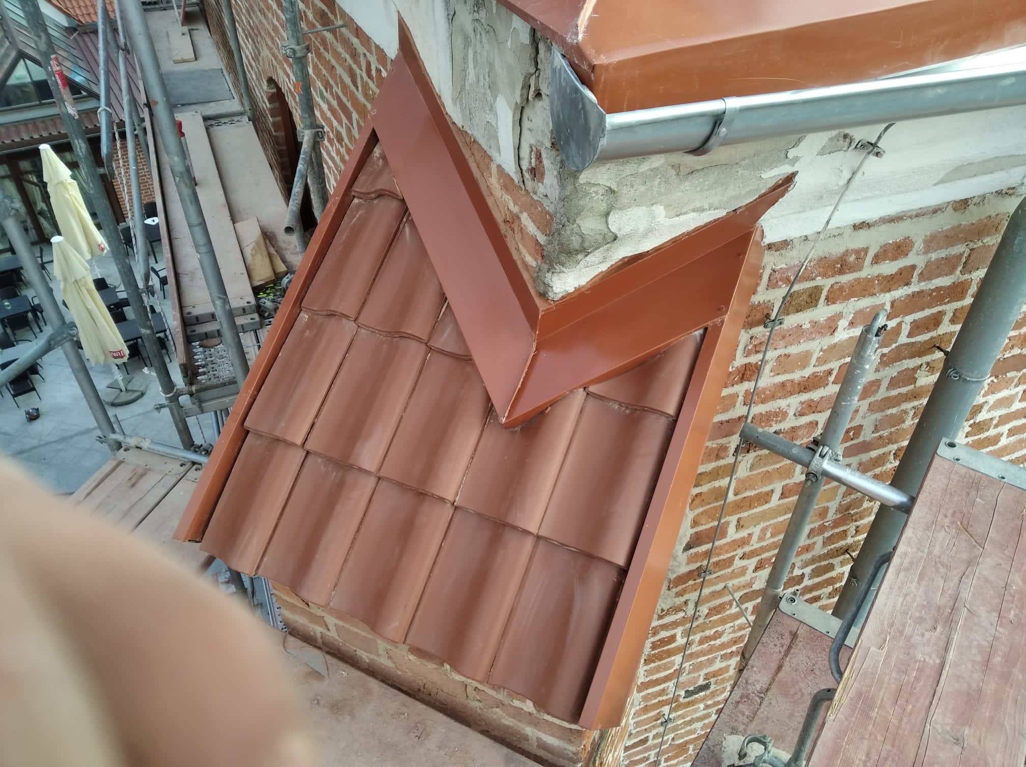 Nowa, brązowa dachówka ceramiczna na murze z czerwonej cegły, z elementami obróbki blacharskiej w kolorze miedzianym, widok z rusztowania.