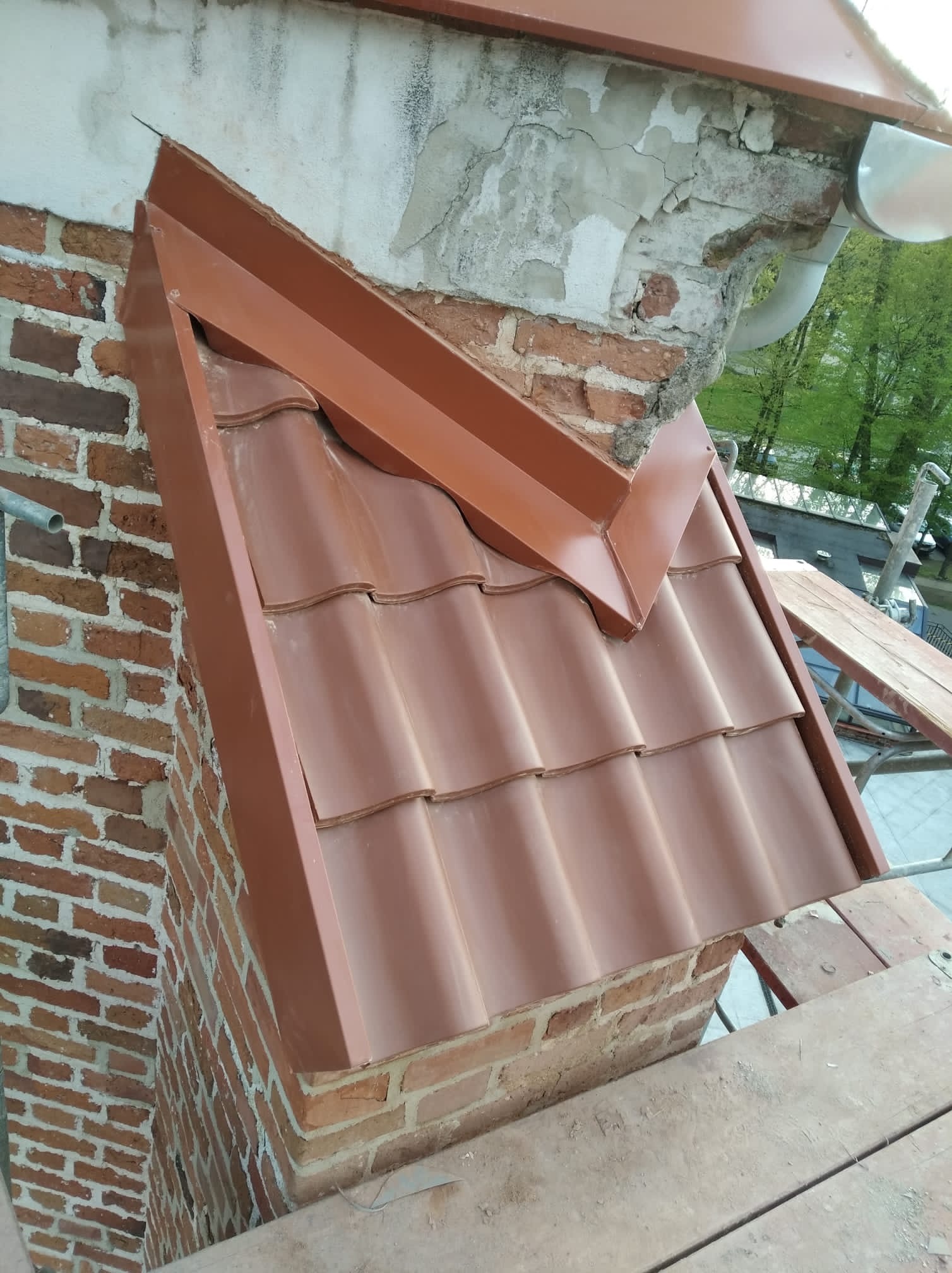 Nowe, brązowe dachówki ceramiczne na kominie z cegły, z obróbkami blacharskimi w kolorze dachówek, widok z rusztowania.