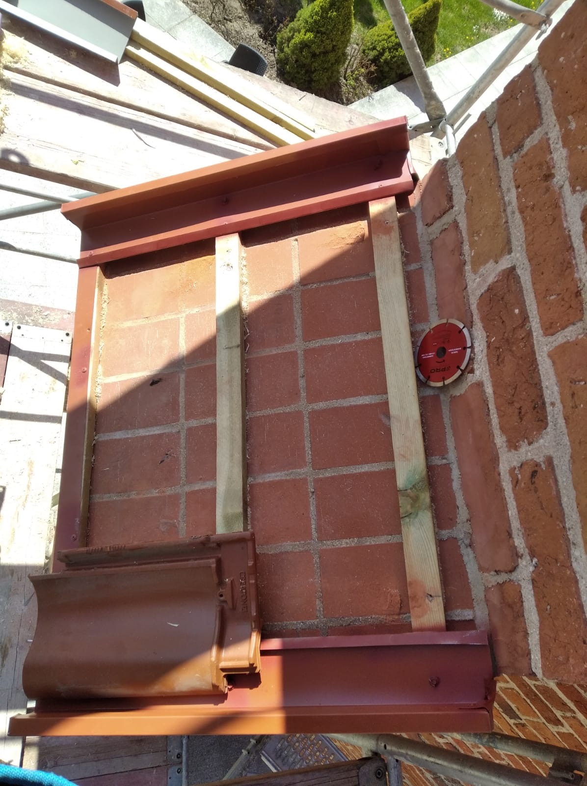 Fragment komina z cegły z zamontowaną metalową ramą, drewnianymi wspornikami i tarczą do cięcia, przygotowany do obróbek blacharskich.