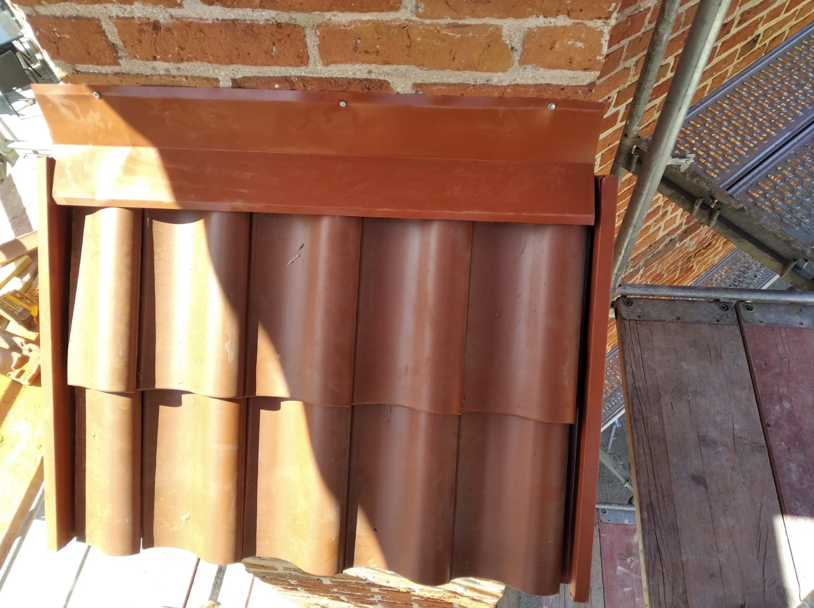 Fragment nowej, brązowej dachówki ceramicznej z obróbką blacharską, zamontowanej na ścianie z cegły, widok z rusztowania.