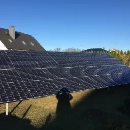 Panele słoneczne ustawione na metalowych wspornikach na trawniku przed domem z ciemnym dachem w słoneczny dzień.