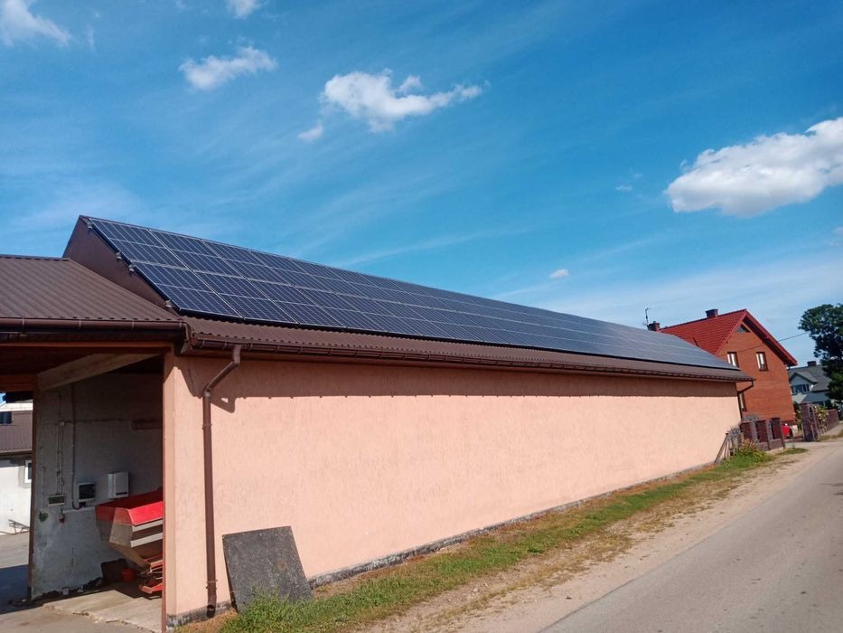 Instalacja paneli słonecznych na dachu budynku gospodarczego z brązowym dachem i jasnoróżową elewacją, widok z poziomu ulicy w słoneczny dzień.