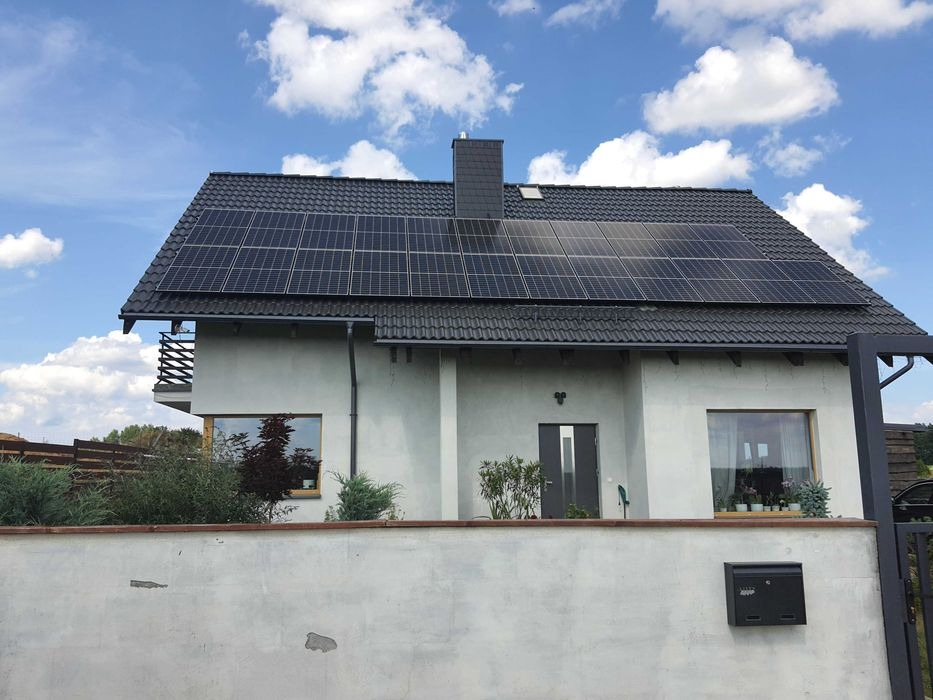 Dom jednorodzinny z panelami słonecznymi na ciemnym dachu, widok zza niskiego muru z czarną skrzynką pocztową, błękitne niebo z chmurami w tle.