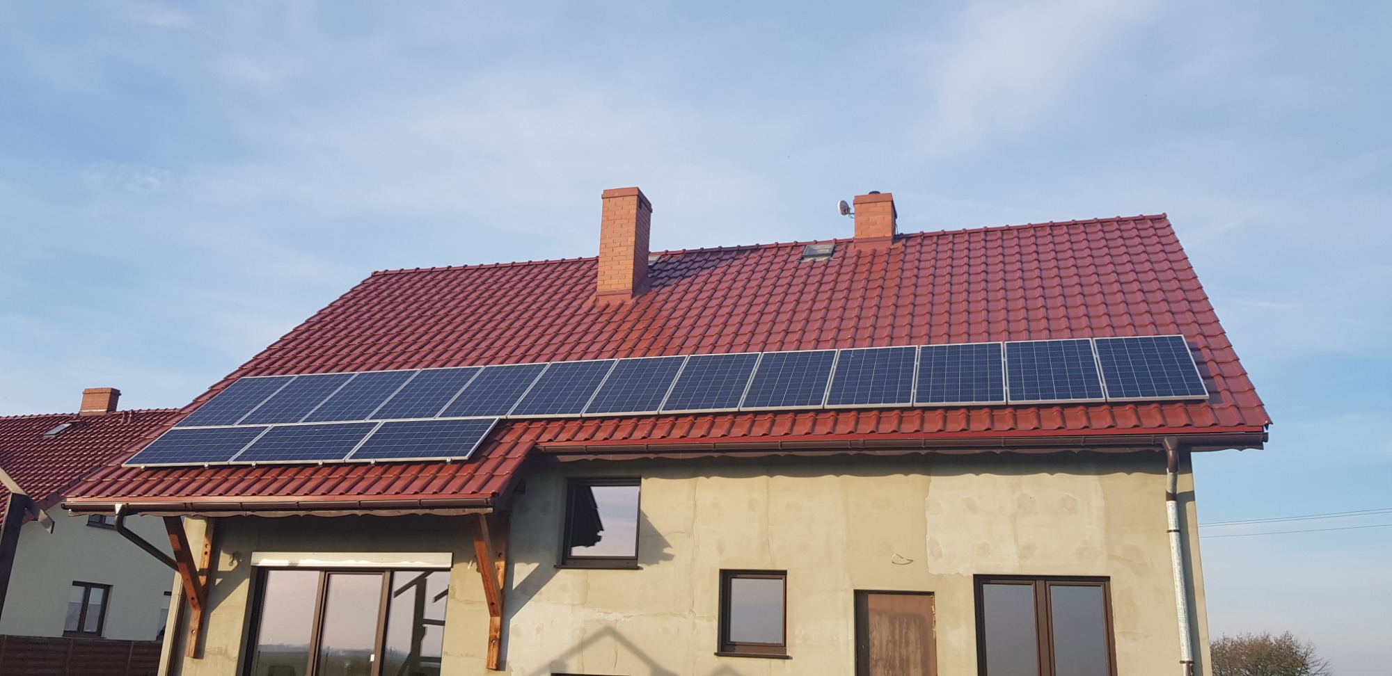 Panele słoneczne zamontowane na czerwonym, spadzistym dachu domu jednorodzinnego z ceglanymi kominami, widoczne okna i fragment elewacji w surowym stanie.