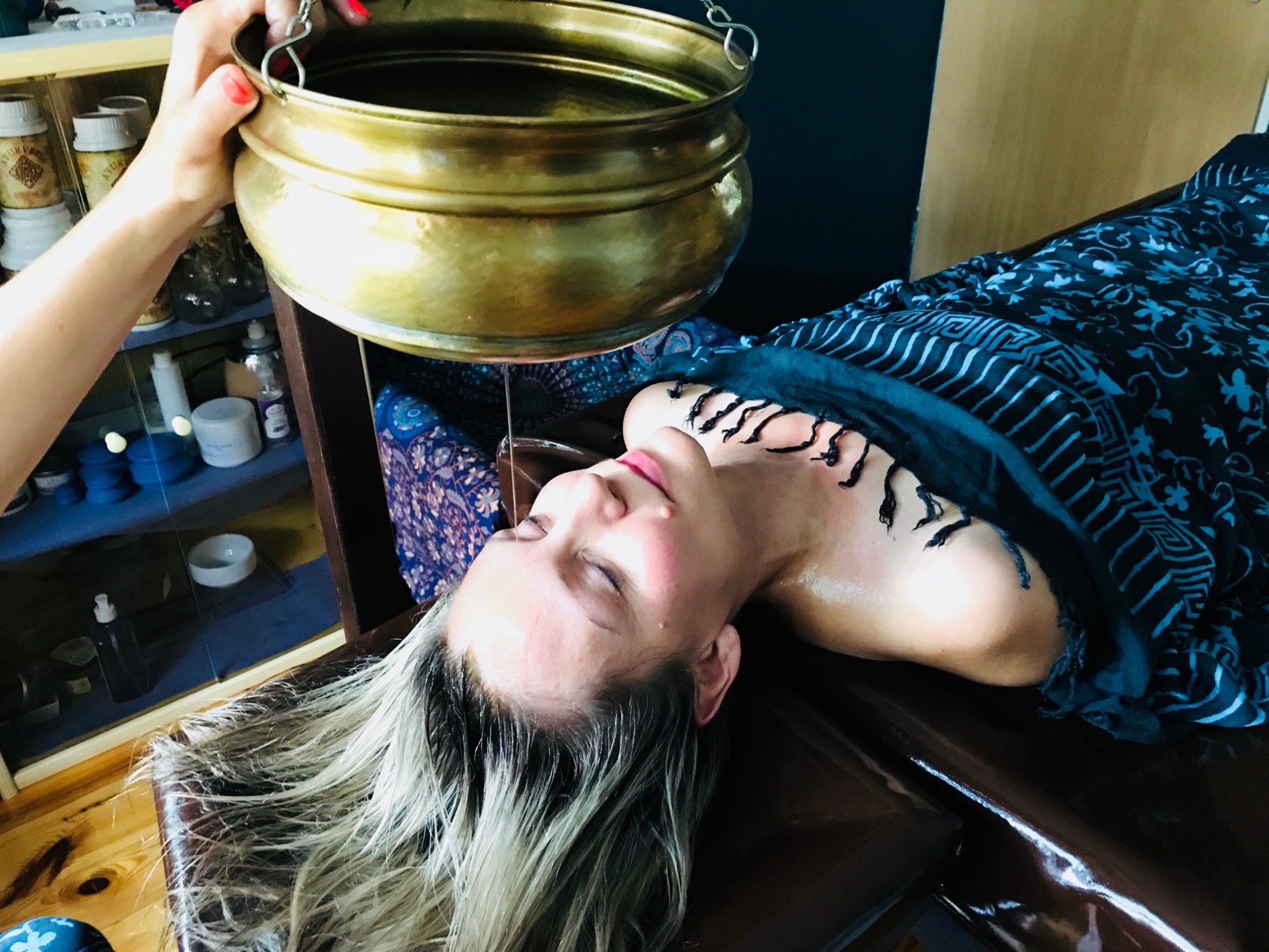 Ajurwedyjski zabieg polegający na polewaniu czoła ciepłym olejem z mosiężnego naczynia zwisającego nad leżącą kobietą, w tle półka z kosmetykami.