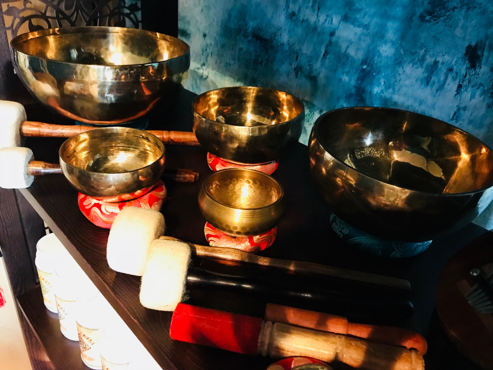 Zestaw złotych mis dźwiękowych różnej wielkości na czerwonych podkładkach, obok pałeczki z białymi i kolorowymi końcówkami, na tle ściany w odcieniach błękitu.