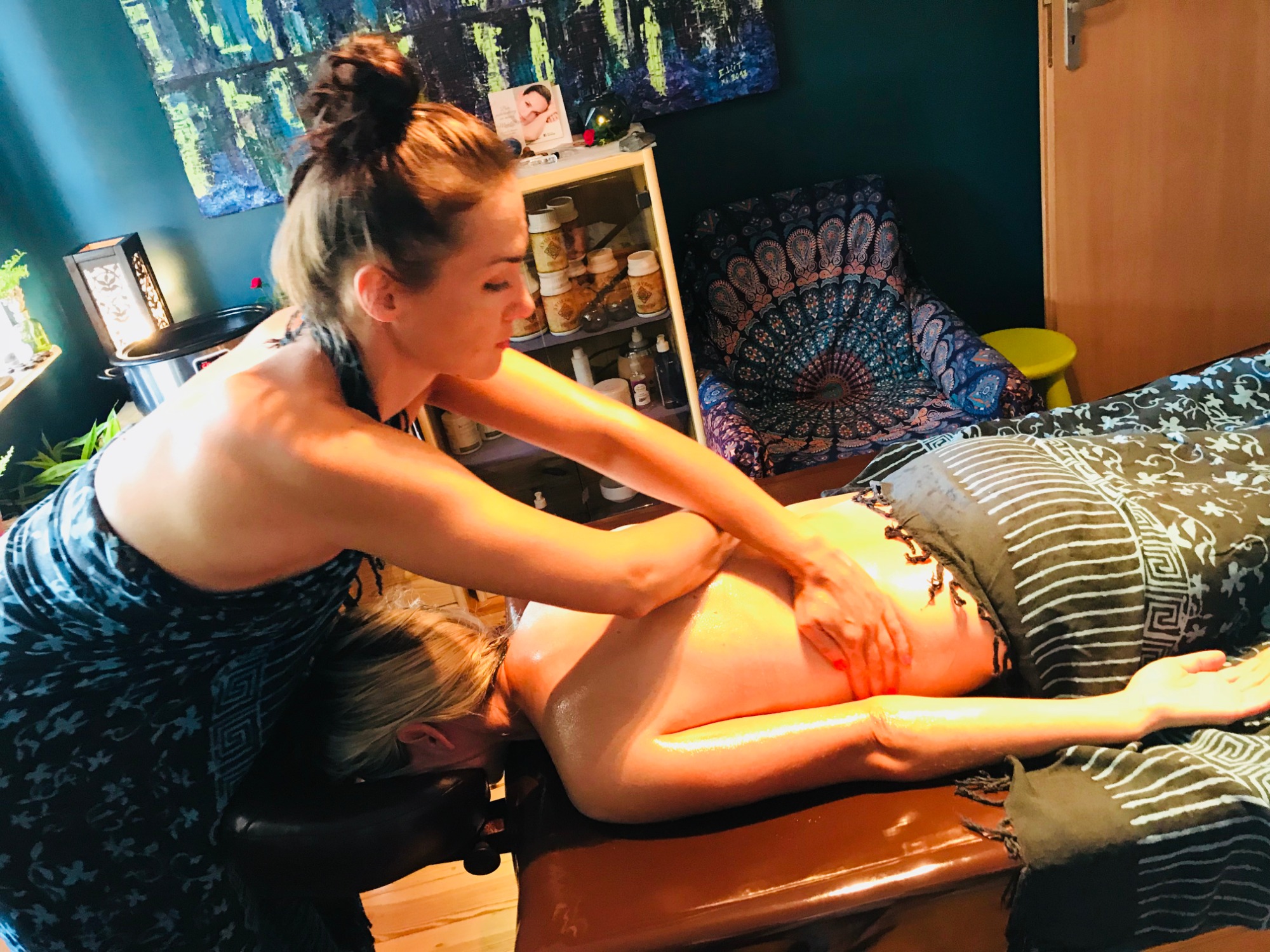 Masażystka w niebieskiej sukience wykonuje masaż pleców klientki leżącej na brązowym stole do masażu, w tle widać szafkę z kosmetykami i dekoracyjny fotel.
