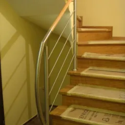 Nierdzewna balustrada z drewnianym poręczą na schodach z zabezpieczeniami kartonowymi na stopniach, żółta ściana w tle.