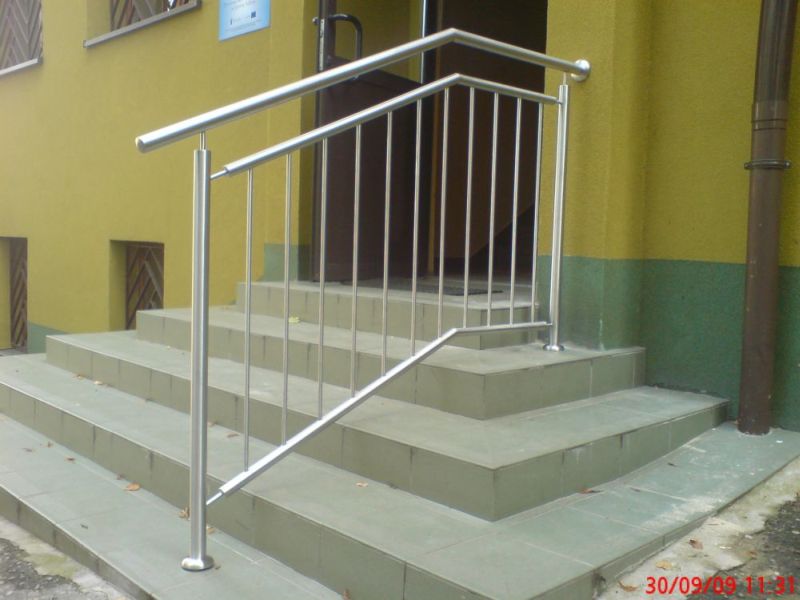 Zewnętrzna, metalowa balustrada ze stali nierdzewnej z pionowymi szczeblami, zamontowana przy schodach wejściowych do budynku z żółto-zieloną elewacją.