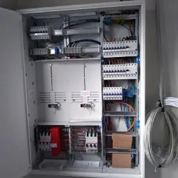 Otwarta skrzynka elektryczna z zamontowanymi bezpiecznikami, przewodami i listwami zaciskowymi, widoczne kable wystające ze ściany.