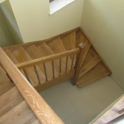 Widok z góry na drewniane schody zabiegowe z drewnianą balustradą z szarymi prętami, prowadzące w dół na wyłożoną kafelkami podłogę, oświetlone przez okno na górze.