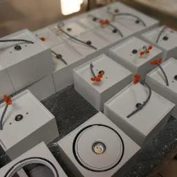 Partia białych, kwadratowych opraw oświetleniowych LED, z wystającymi przewodami i pomarańczowymi złączkami, ułożonych na folii bąbelkowej, jeden z opraw jest zmontowany i pokazuje gotową lampę.