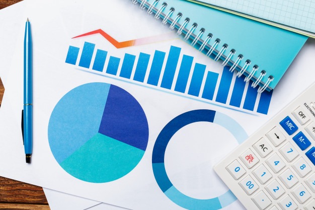 Analiza danych finansowych: wykresy kołowe i słupkowe w odcieniach niebieskiego, kalkulator, notatnik i długopis na drewnianym biurku.