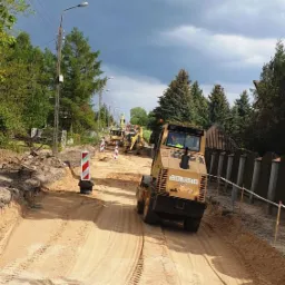 Żółty walec drogowy BOMAG ubija piasek na drodze, w tle koparki i słupki ostrzegawcze, pochmurne niebo.