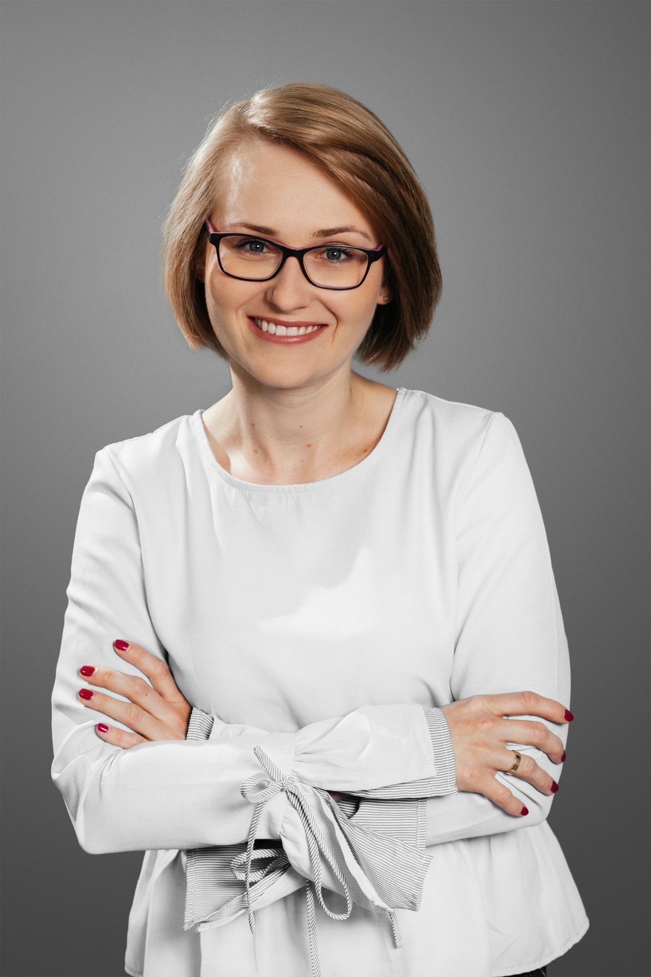 Portret uśmiechniętej kobiety w okularach, z ramionami skrzyżowanymi na piersiach, w białej bluzce z ozdobnymi wiązaniami na rękawach, na szarym tle.