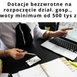Kobieta w białej koszuli i czarnym krawacie wskazuje palcem na plan architektoniczny wyświetlany na ekranie laptopa, obok smartfon z tym samym planem, notatnik i długopis na biurku, informacja...