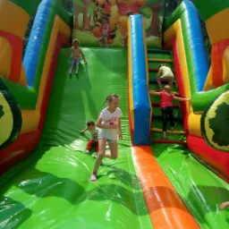 Dzieci bawiące się na dmuchanej zjeżdżalni z motywem Kubusia Puchatka, dziewczynka w białej koszulce biegnie na dół, inne dzieci wspinają się po schodach i zjeżdżają.