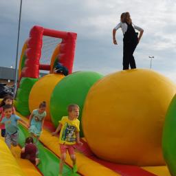 Jupilandia Organizacja Eventów dla Dzieci i Dorosłych - Dzieci bawią się na dmuchanym torze przeszkód z dużymi, kolorowymi kulami i siatką, na tle budynku sklepowego i pochmurnego nieba.