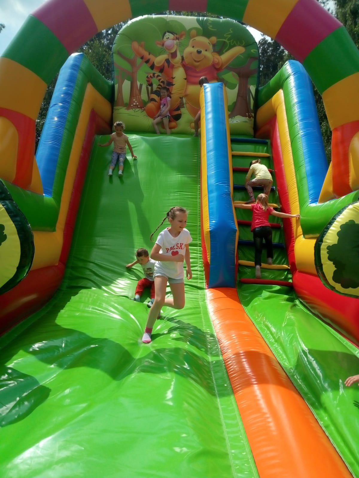 Dzieci bawiące się na dmuchanej zjeżdżalni z motywem Kubusia Puchatka, dziewczynka w białej koszulce biegnie na dół, inne dzieci wspinają się po schodach i zjeżdżają.