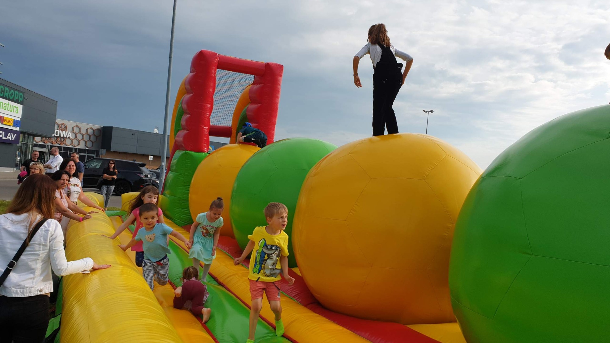 Dzieci bawią się na dmuchanym torze przeszkód z dużymi, kolorowymi kulami i siatką, na tle budynku sklepowego i pochmurnego nieba.