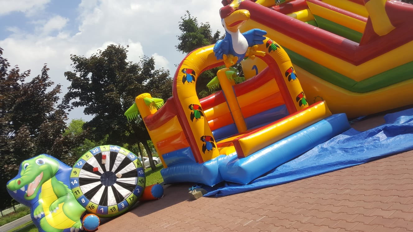 Kolorowy dmuchany zamek z motywem dżungli, z figurą tukana na górze oraz dmuchana tarcza do darta z krokodylem, ustawione na kostce brukowej, częściowo przykryte niebieską plandeką.