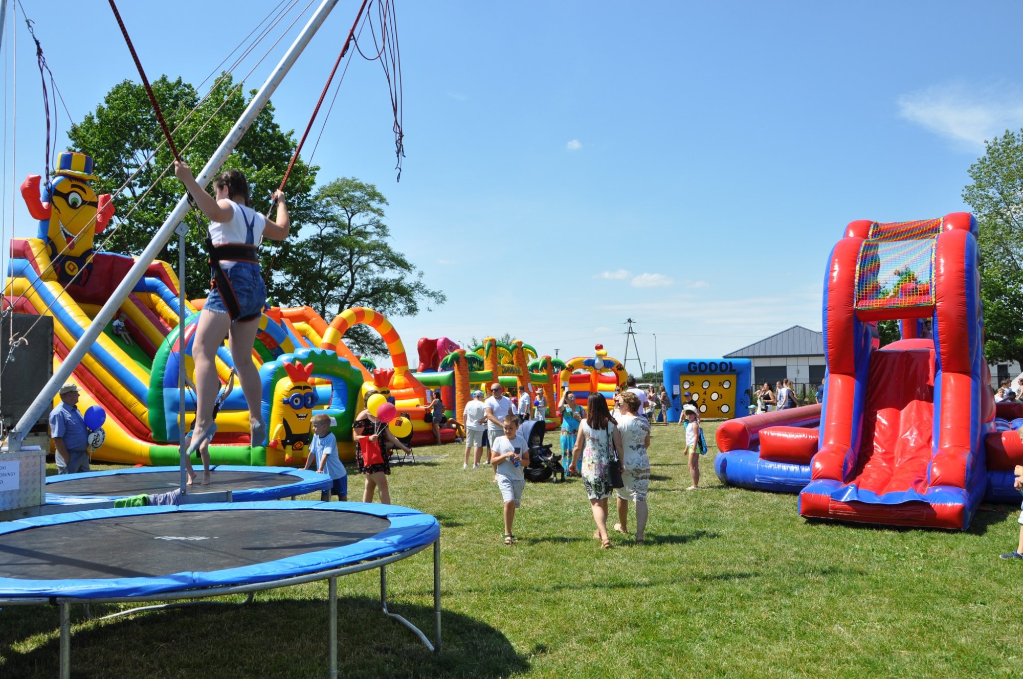 Dziewczynka w uprzęży skacze na trampolinie bungee na tle dmuchanych zamków i bawiących się ludzi podczas imprezy plenerowej.