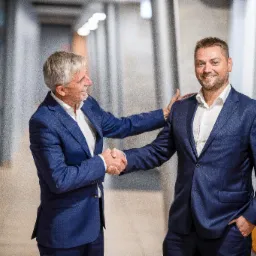 Dwóch mężczyzn w garniturach w jasnym korytarzu uśmiechniętych, wymieniających uścisk dłoni, jeden z nich kładzie dłoń na ramieniu drugiego.