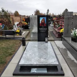 Granitowy nagrobek z portretem dziecka i motywem skrzydła, otoczony innymi nagrobkami na cmentarzu.