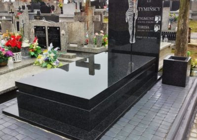 Czarny, polerowany nagrobek z granitu z wygrawerowanym wizerunkiem Chrystusa i zdjęciami zmarłych, ustawiony na cmentarzu.