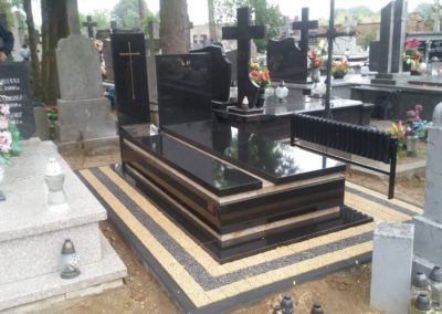 Czarny, warstwowy pomnik z krzyżem na cmentarzu, otoczony kostką brukową w odcieniach beżu i czerni, w tle inne nagrobki i drzewa.