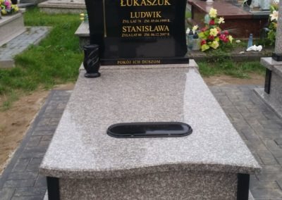 Granitowy pomnik z czarną tablicą nagrobną, inskrypcjami i wazonem, umieszczony na cmentarzu z kostką brukową.