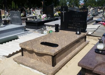 Jasny, granitowy grobowiec z czarnym krzyżem i inskrypcją na cmentarzu, otoczony innymi nagrobkami i zielenią drzew.