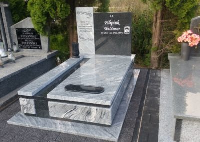 Szary, podwójny pomnik z czarnym nagrobkiem z fotografią osoby zmarłej, otoczony innymi nagrobkami i zielenią.
