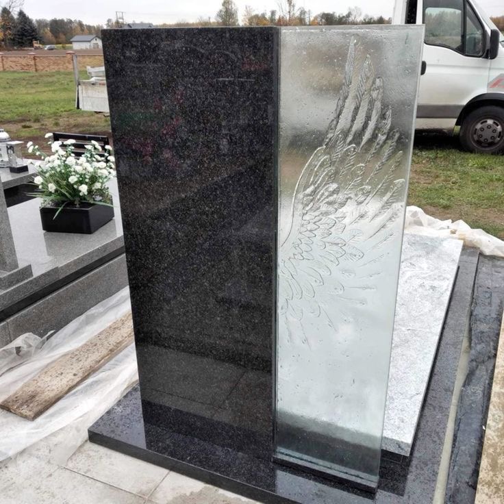Dwuczęściowy pomnik z ciemnego granitu i przezroczystego szkła z wygrawerowanym motywem skrzydła, ustawiony na płycie granitowej.